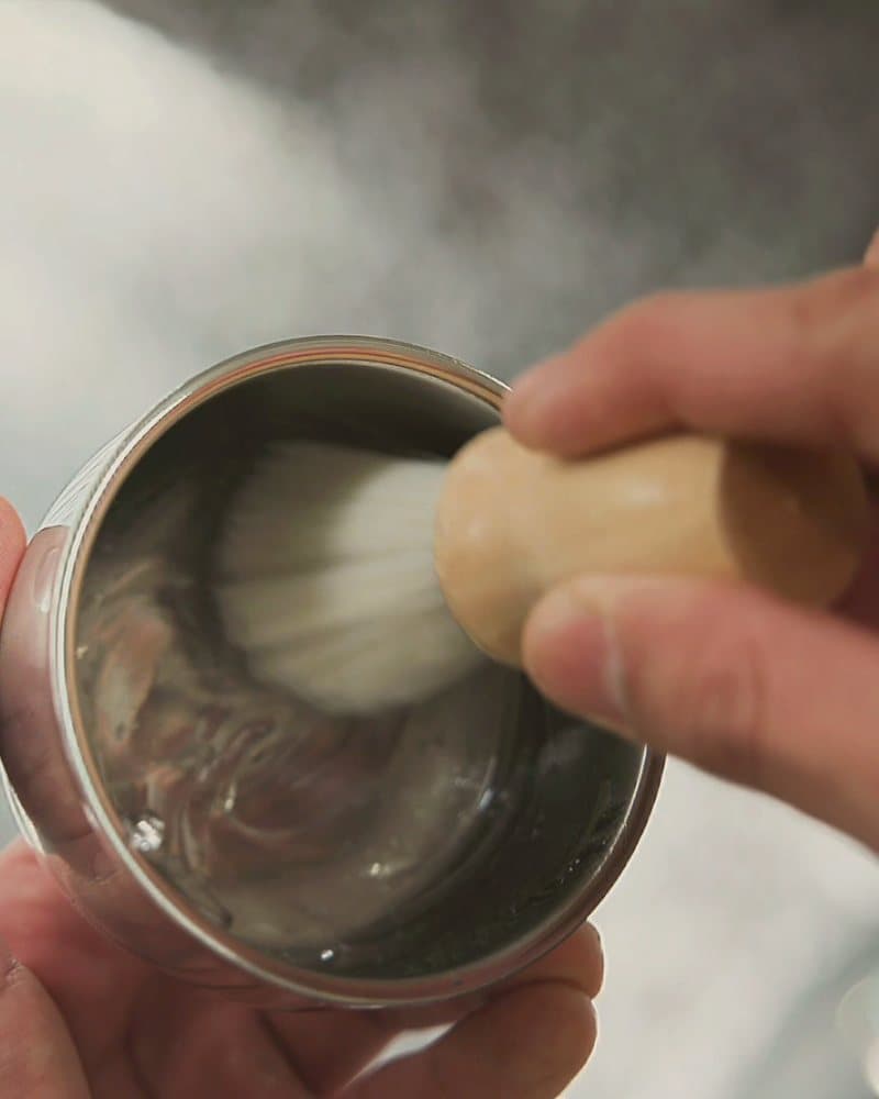 Manos de barbero montando la espuma con brocha y cuenco de acero.