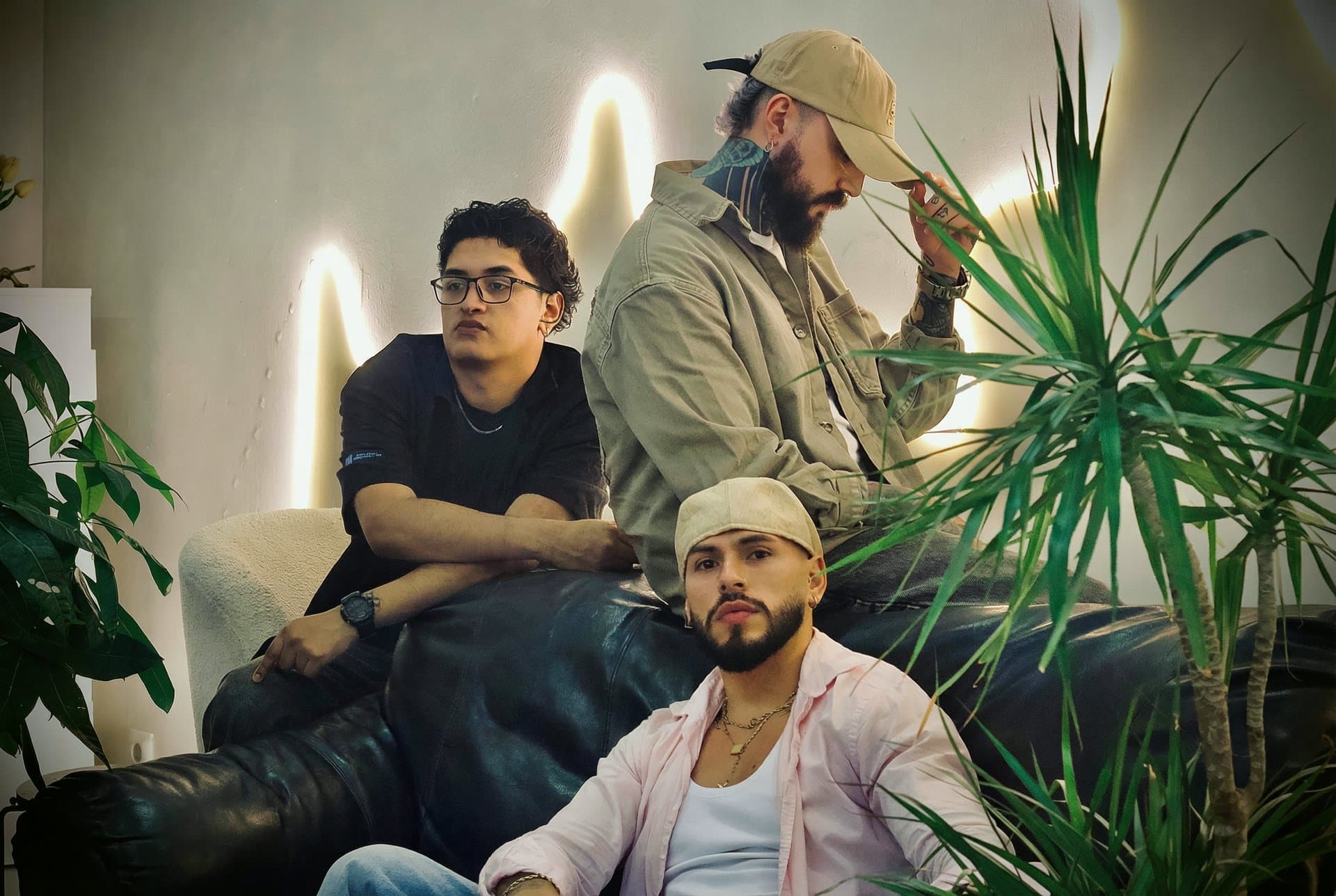 Tres barberos en el sofá de cuero de una barbería, entre plantas y con la pared iluminada detrás.